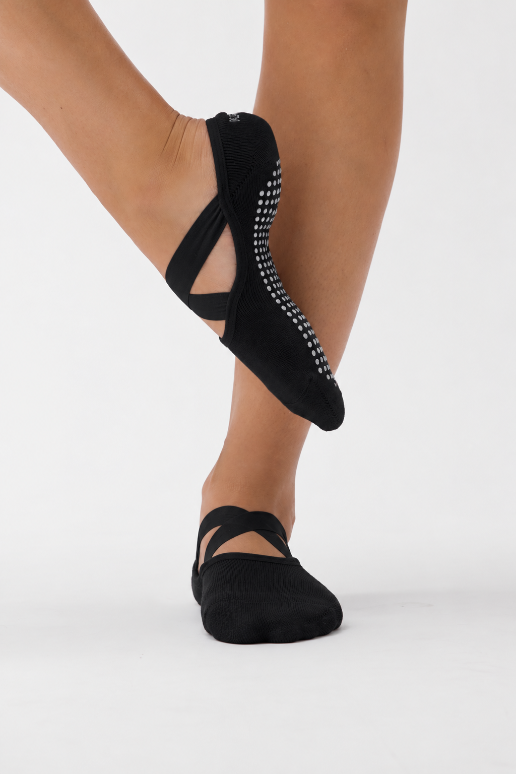 Black pilates grip socks with crisscross ankle straps and a black-and-white dotted grip sole, shown on a dancer's feet against a light gray studio background.