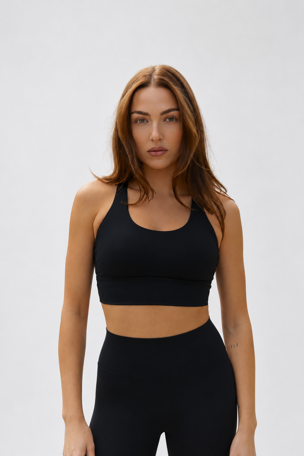 A woman modeling a black sports bra and matching black high-waisted leggings against a light gray studio background.