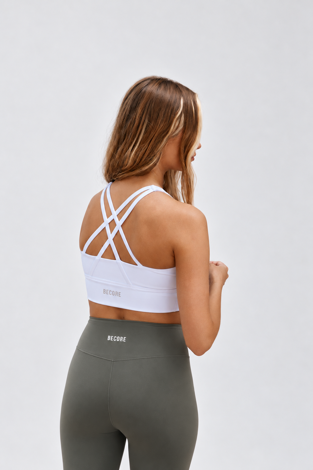 A woman shown from the back wearing a white sports bra with crisscross straps and olive green leggings, photographed against a plain light gray studio background.