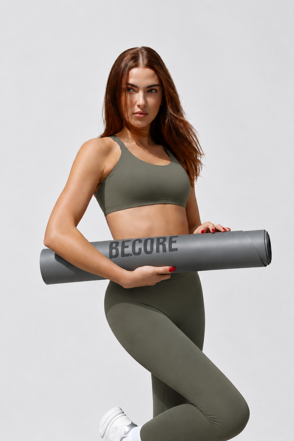 A woman in an olive green sports bra and matching leggings holds a gray yoga mat with the brand name BECORE printed on it, posed against a light gray studio background.