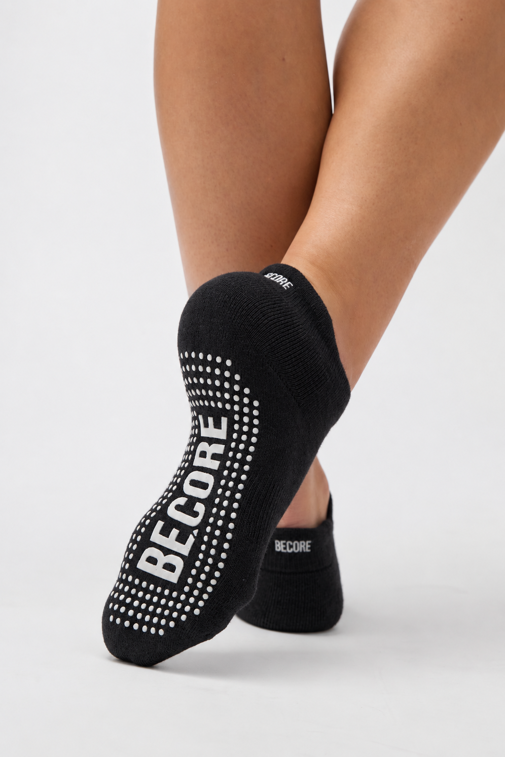 Black low-cut ankle socks with white grip dots and the brand name BECORE printed on the sole, shown on a model's feet against a light gray studio background.